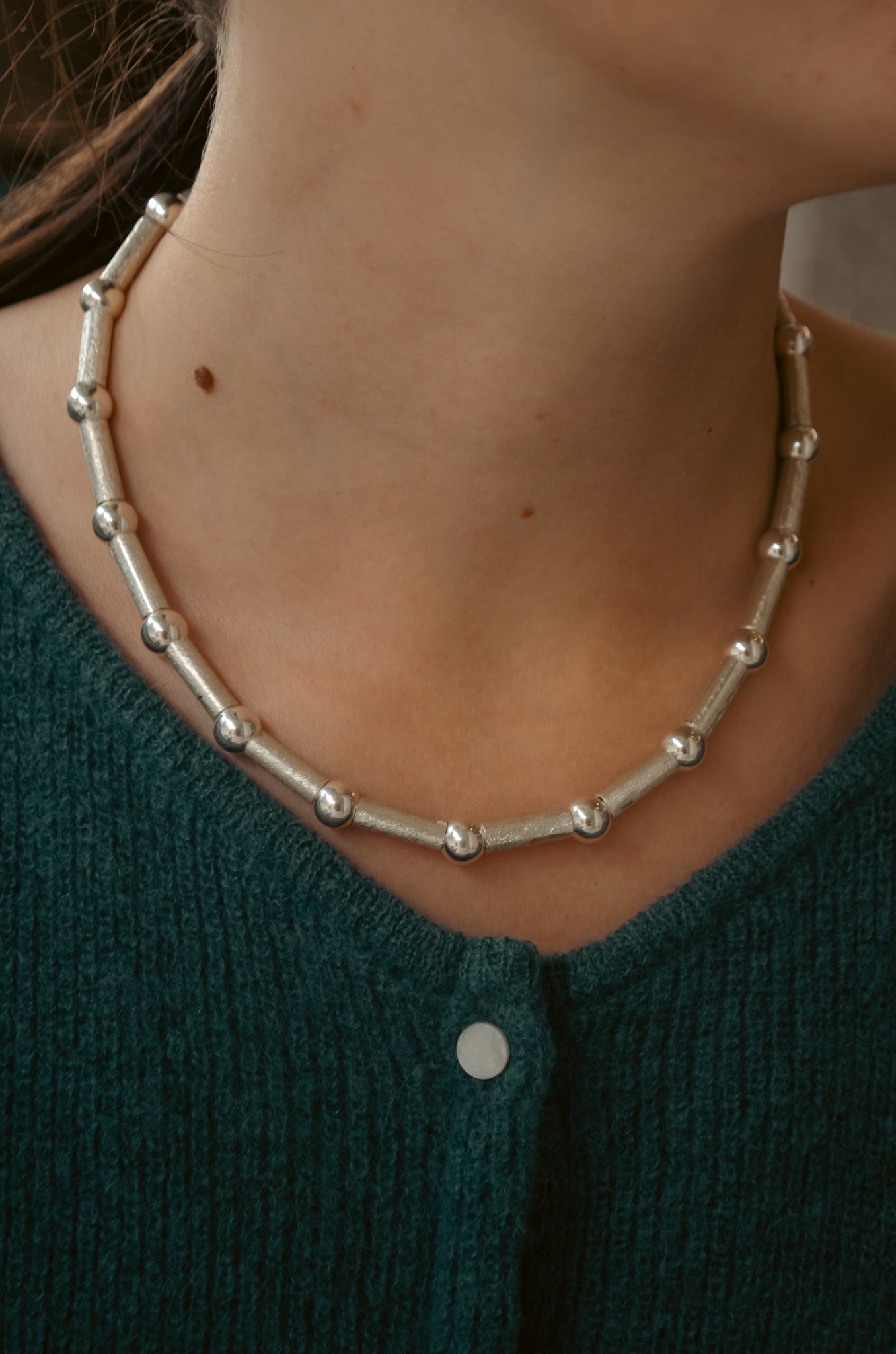 Close-up of a silver necklace alternating scratch-textured tubes with round polished beads worn by woman in dark green knit sweater.